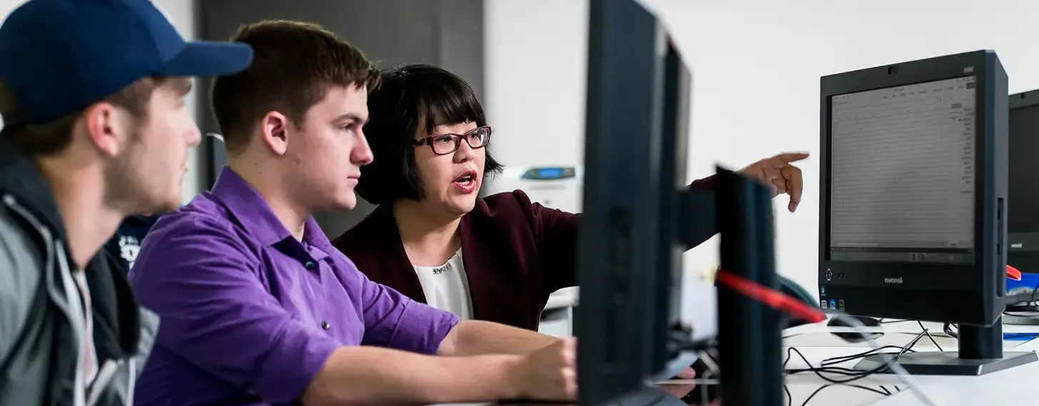 Students focused on computers in a classroom with their professor pointing something out on the screen.